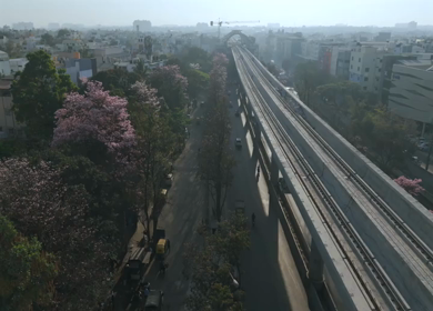 Aerial Slow Motion View of Metro Line and Road at Silk Board Junction with Pink Blossom Trees Bengaluru